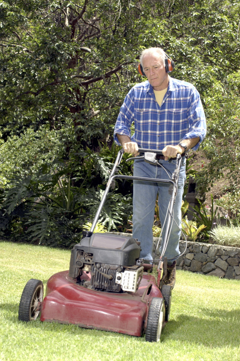 A man wearing ear protection pushes a lawn mower across the grass toward the viewer.