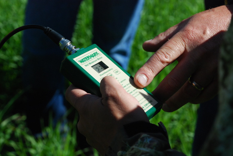 Some gardeners using a soil moisture meter, with a finger pointing to the readouts.